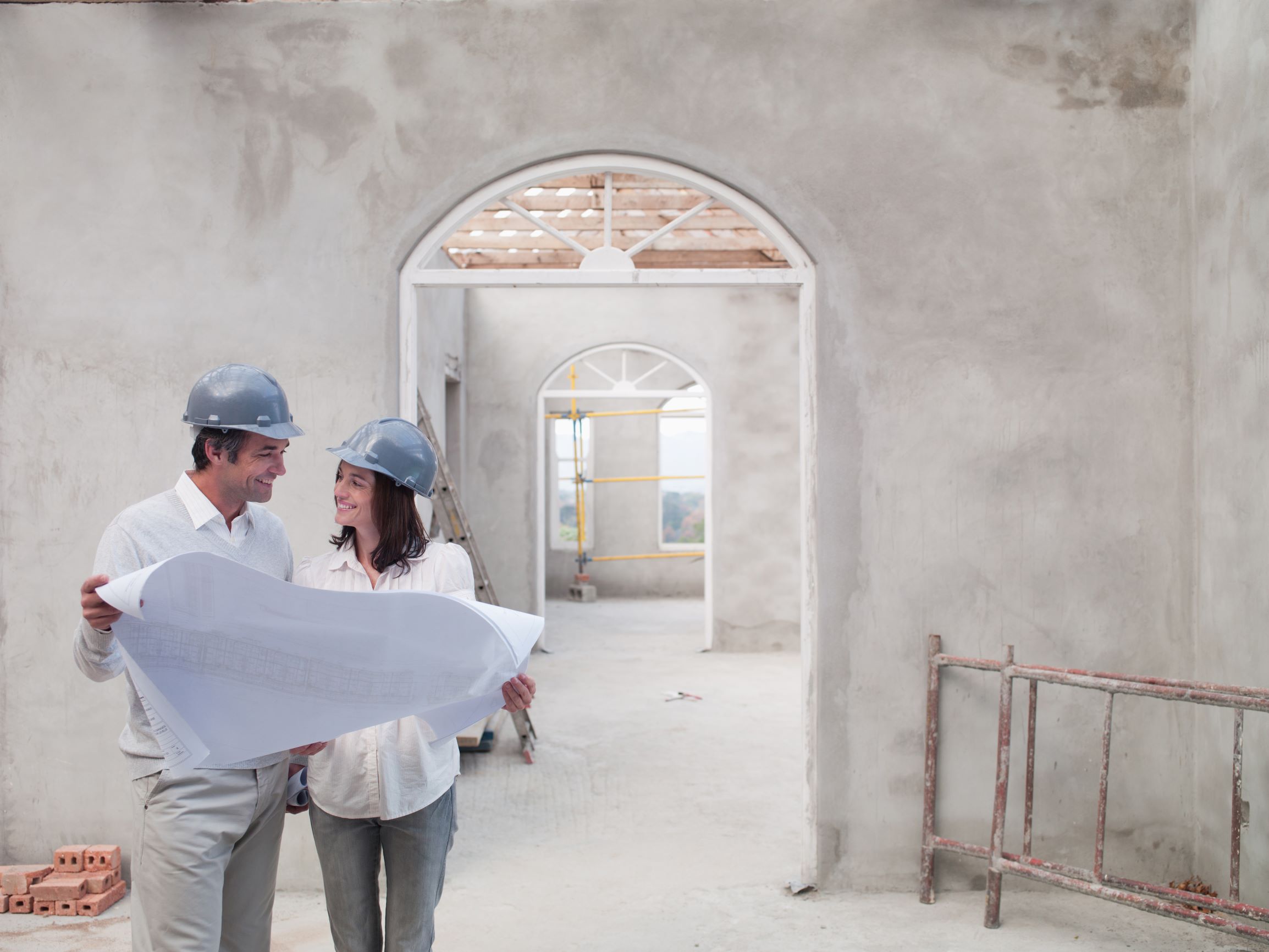 Homme et femme souriants portant des casques et tenant un plan de construction. Le couple se trouve dans un bâtiment en train d'être construit. 
